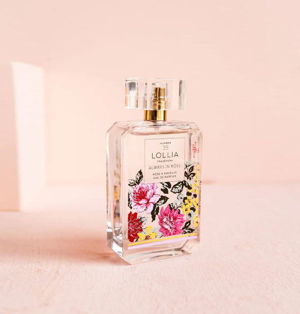 Clear glass bottle of Lollia Always In Rose Eau de Parfum with beveled accents, colorful floral design, and faceted cap over a gold nozzle