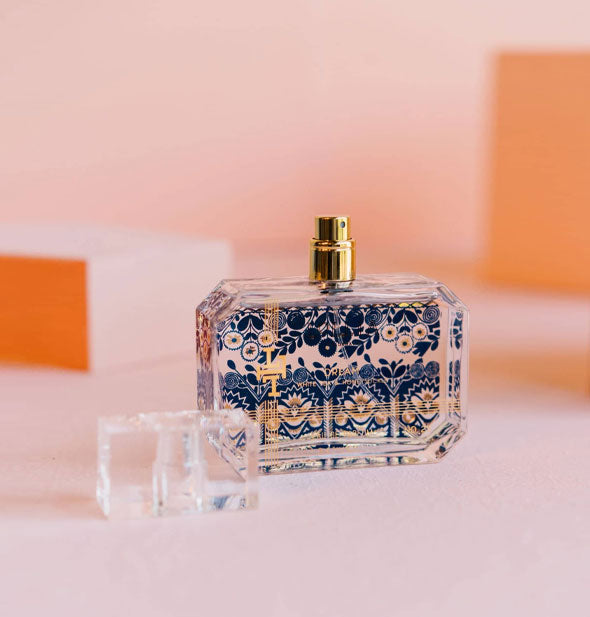 Rectangular glass bottle with beveled corners, gold spray nozzle, and blue floral designs with gold accents on a pink backdrop