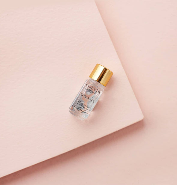 A small glass bottle of Lollia Elegance Eau de Parfum with gold cap rests on a rectangular pink platform