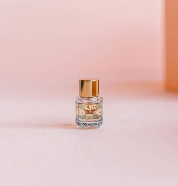 Cylindrical clear glass mini bottle of Lollia fragrance is topped with a gold cap and sits against a pink backdrop