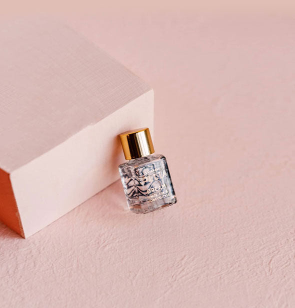 Small glass Lolla Dream Eau de Parfum bottle with blue florals and a gold cap is propped against a rectangular pink platform on a textured pink surface