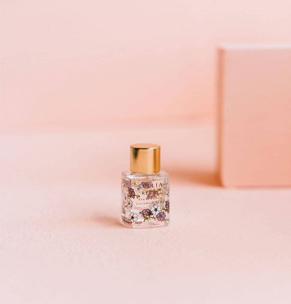 Small rectangular glass bottle of Lollia fragrance with all-over floral designs and a gold cap sits against a pink backdrop