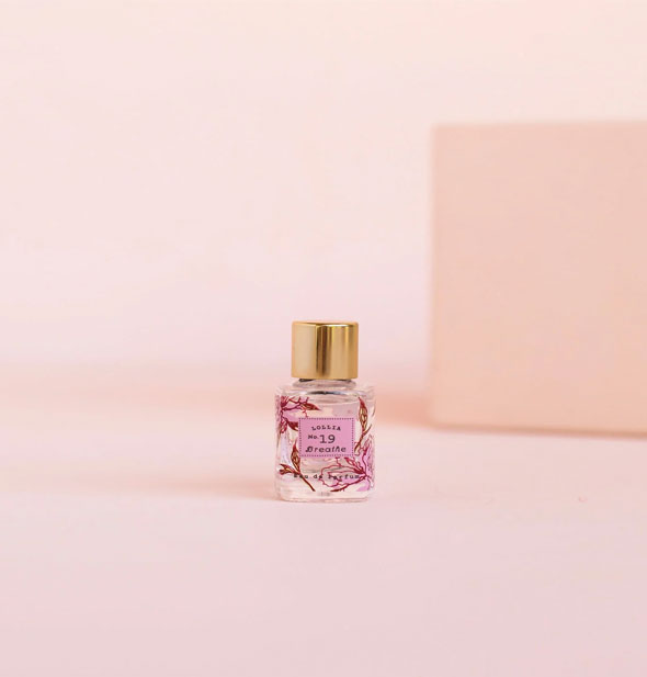 Small glass bottle of Lollia No. 19 Breathe perfume against a pink backdrop features a pink floral design and gold cap