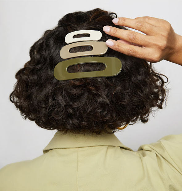 Model wears three hair clips—one white, one tan, and one green—with an elongated oval shape and center opening in a partially pulled-back hairstyle