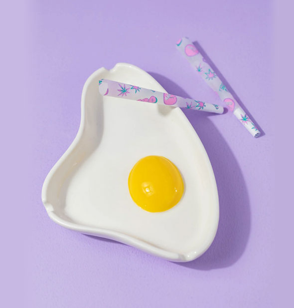 White ashtray with raised yellow "yolk" in the center resembles a fried egg and is staged with two rolled cigarettes against a purple backdrop