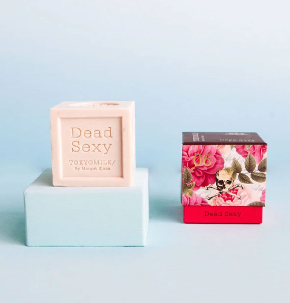 Square bar of Dead Sexy TokyoMilk soap sits on a blue platform next to its box packaging decorated with pink florals, a red base, and skull and crossbones accent