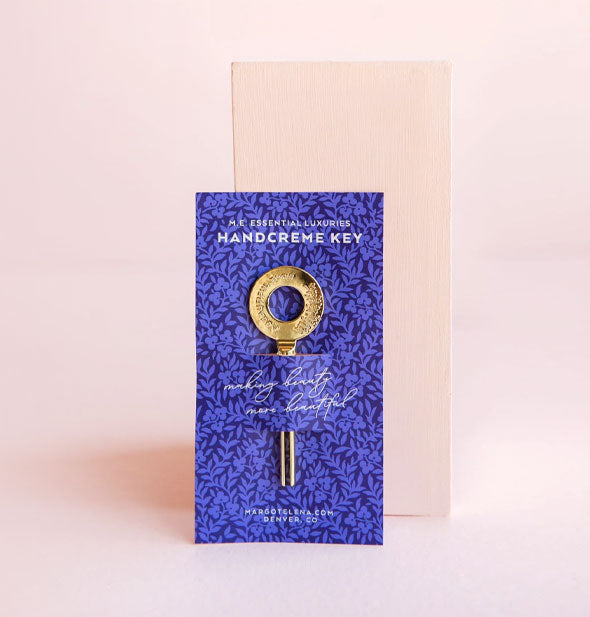 Gold Essential Luxuries Handcreme Key on purple floral patterned product card in front of a pink block backdrop