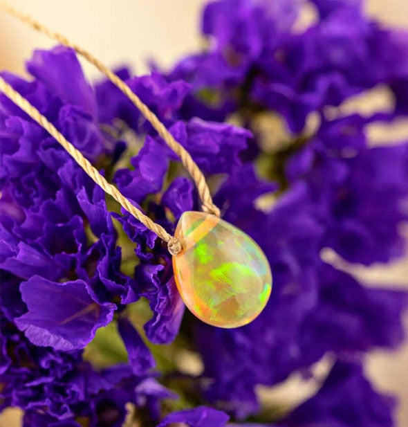 A green iridescent teardrop-shaped Ethiopian opal stone necklace on gold cord rests on purple flower petals