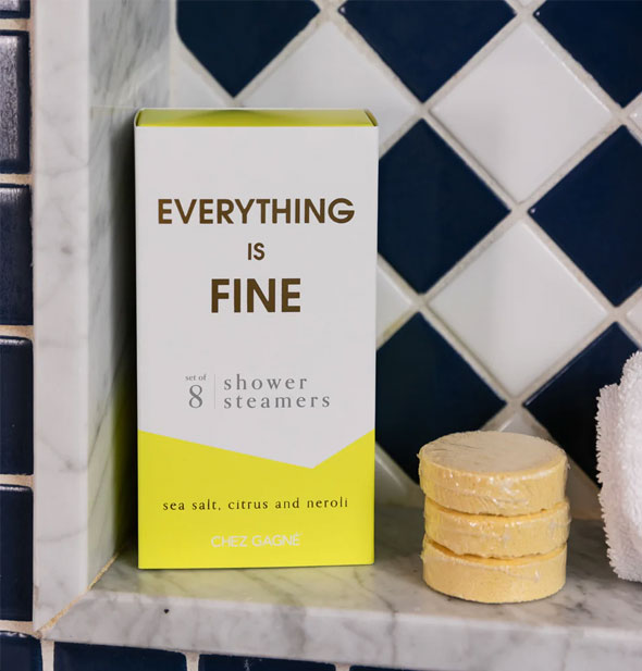 Everything Is Fine shower steamers box and three yellow steamers rest on a marble bath ledge next to a rolled-up white washcloth in front of black and white tile