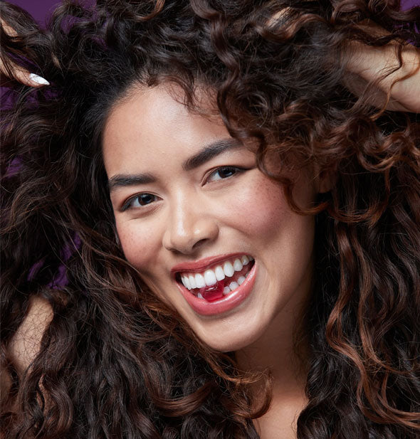 Smiling model with hands in her full, healthy-looking curly hair holds a vitamin gummy between her teeth