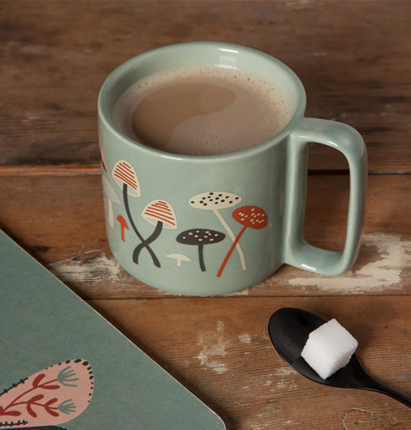 Far and Away Mushroom Midi mug is filled with a slightly foamy coffee beverage and sits on a wooden tabletop with black spoon holding a sugar cube and a matching green notebook partially visible
