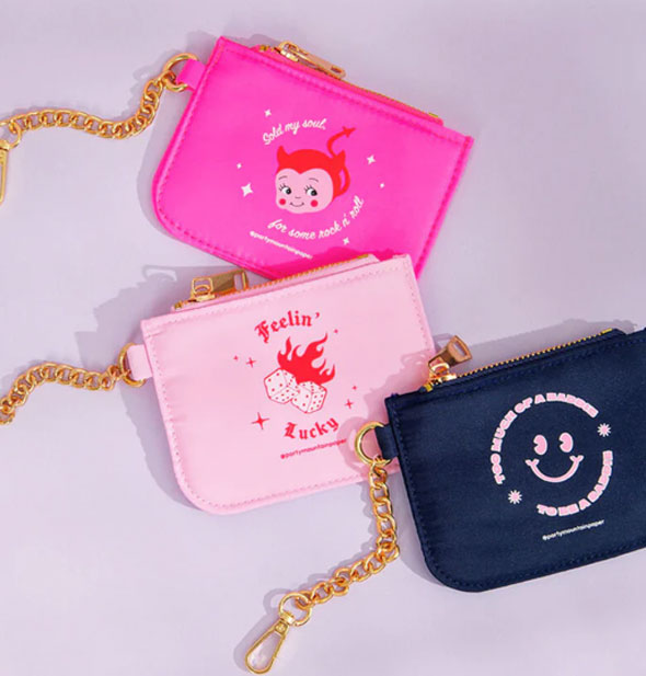 Three styles of coin purses with gold chains attached: navy blue Baddie/Saddie smiley, light pink Feelin' Lucky dice, and hot pink Sold My Soul devil