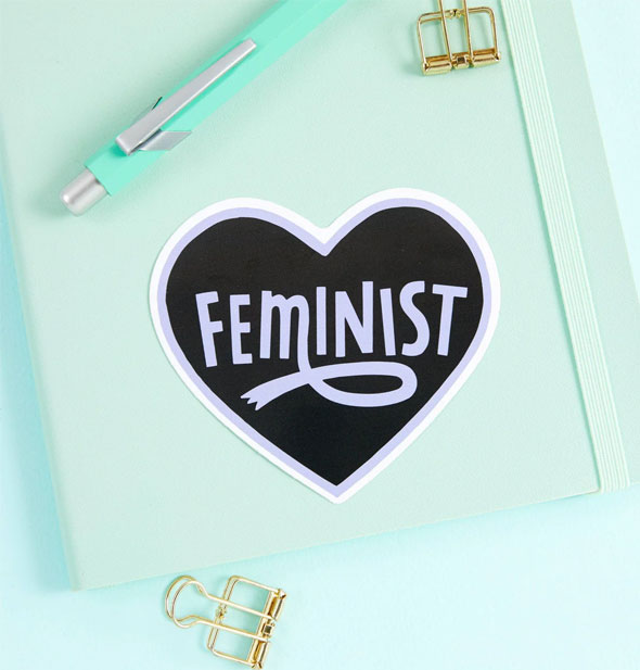 Feminist heart sticker on a green notebook cover with pen and gold paperclips to the side