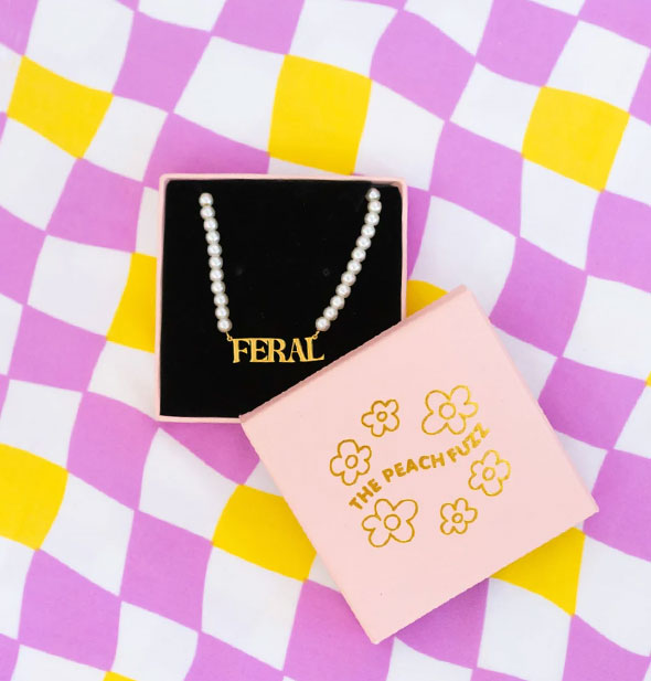 Gold "FERAL" nameplate necklace on a string of pearls in an opened The Peach Fuzz gift box on a wavy purple, white, and yellow checkered backdrop