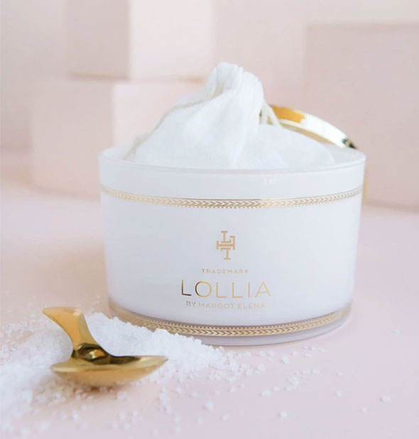 Opened Lollia container reveals a white drawstring bag inside; bath salts are strewn in front with a gold spoon resting on top