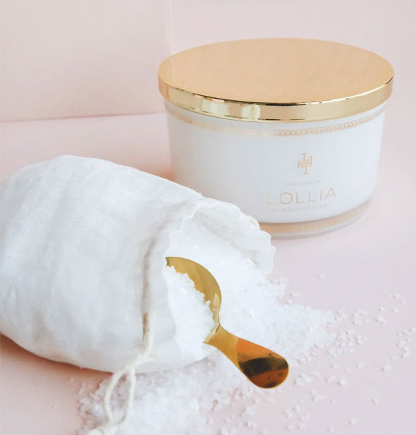 White and gold Lollia tub sits in the background of an opened white drawstring bag with bath salts spilling out around a gold spoon