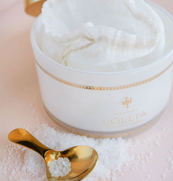 An opened tub of Lollia bath salts reveals a white drawstring bag inside. Salts are scattered in front around and in a gold spoon