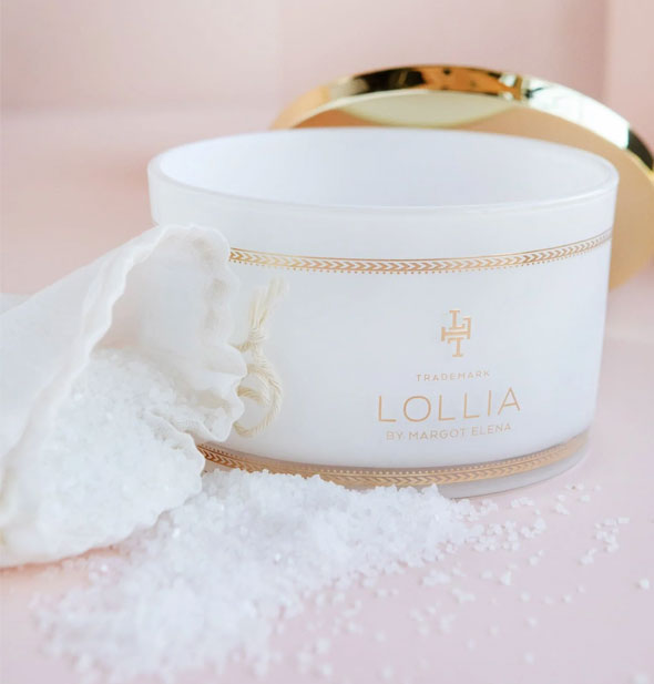An opened white container of Lollia bath salts with shiny gold lid set behind it and salts strewn about in the foreground, spilling out of a white drawstring bag
