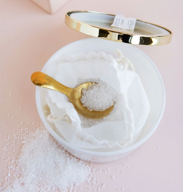 Top view of an opened tub of Lollia bath salts with shiny gold lid placed behind, salt crystals strewn about, and a gold spoon dipped into the salt-filled white drawstring bag inside