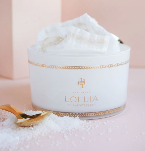 Opened tub of Lollia bath salts with white drawstring bag inside. Salts are piled and scattered in front with a gold spoon