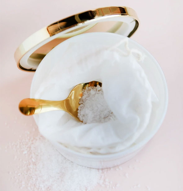 Top view of an opened tub of bath salts with shiny gold lid removed to reveal a white drawstring bag inside with gold spoon and some salt crystals spilled out