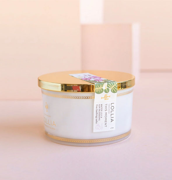 Whitish tub of Lollia bath salts with shiny gold lid secured by a floral sticker label