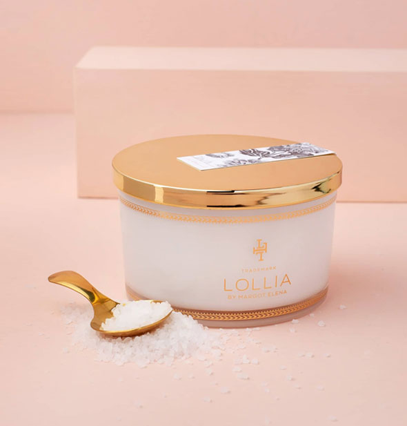 Jar of Lollia bath salts with gold lid and spoon, in and around which is scattered salt granules