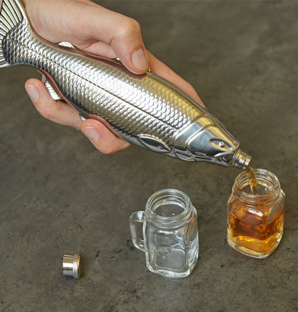 Model pours brown liquor from a fish flask into a small glass jar