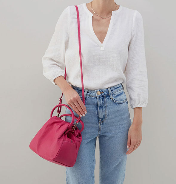 Model wearing jeans and a white shirt wears a fuchsia-colored leather satchel bag over shoulder with an elongated strap