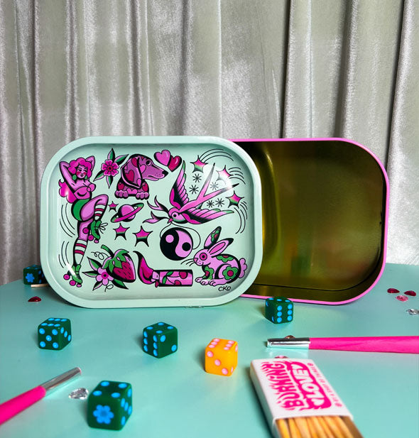 Rectangular metal box with pink rim features a lid with pink tattoo art that doubles as the box's lid. It rests on a mint green surface with colorful dice, matches, and pink cigarettes