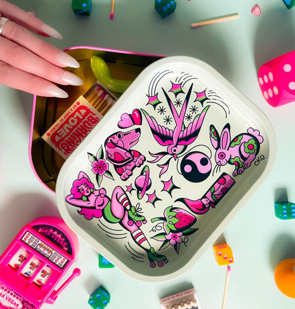 Model's hand touches the rim of a partially-opened flash art box with rolling tray lid that is filled and surrounded with random items
