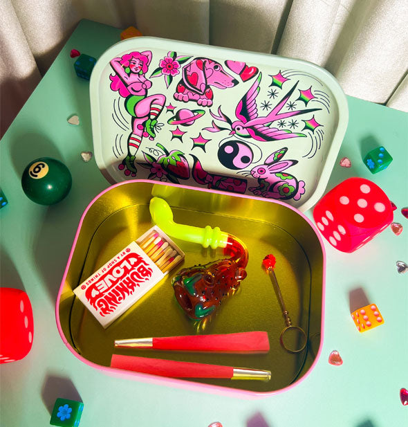 Rectangular metal box contains smoking items; its flash art tray lid rests upright behind it, both surrounded by colorful dice and pool balls