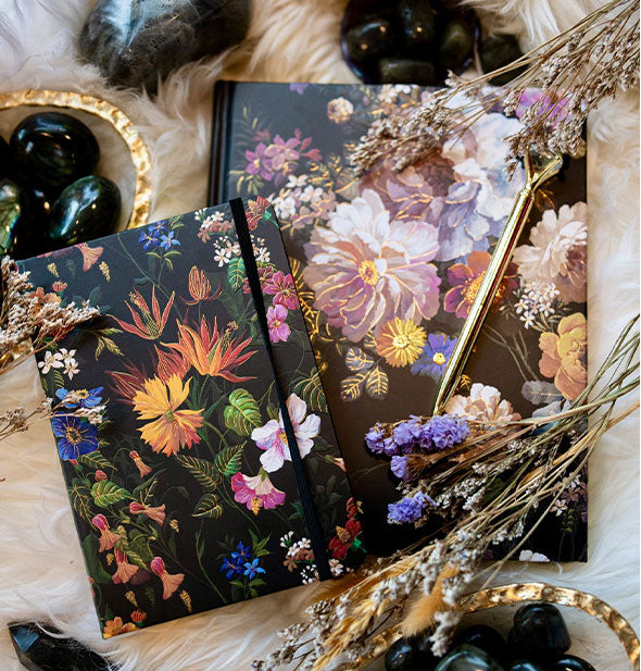 Two black journals with colorful floral patterns rest on a fur surface scattered with dried flowers and black gemstones