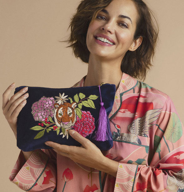 Smiling model poses with a dark blue velvet pouch featuring colorful floral and tiger head embroidery design and a purple tassel zipper pull