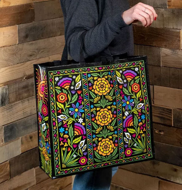 Model wears the straps of the Flower Fest Shopper over one arm against a wooden backdrop