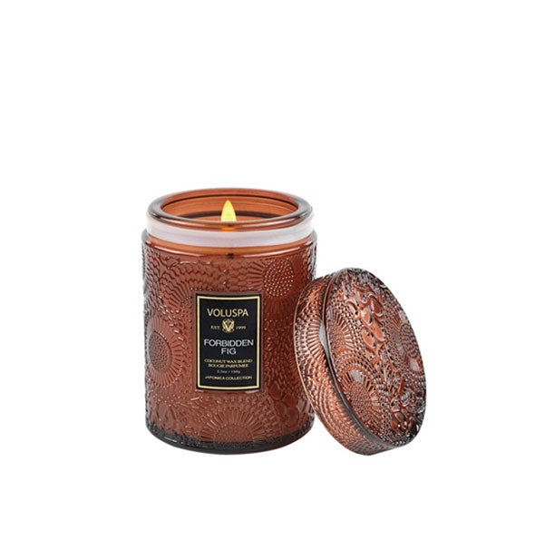 Brown embossed glass candle jar with matching glass lid propped against it features a lit wick and black Voluspa: Forbidden Fig label