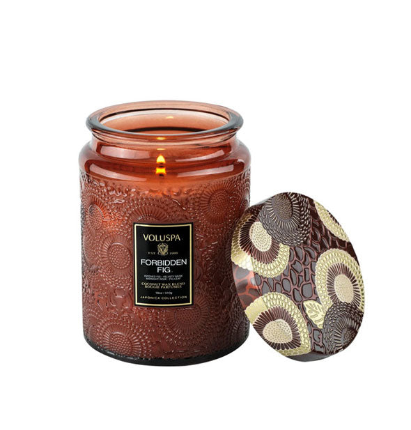 Brown embossed glass candle jar with ornately decorated metal lid propped against it features a lit wick and black Voluspa: Forbidden Fig label