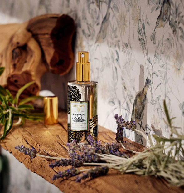 A bottle of Voluspa French Cade Lavender Room & Body Mist rests on a rustic wooden shelf staged with dried flowers and driftwood against delicate bird and flower patterned wallpaper