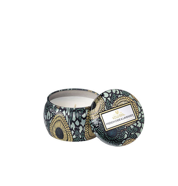 A small unlit candle inside a rounded tin with blue and gold metallic floral design and matching lid set to the side