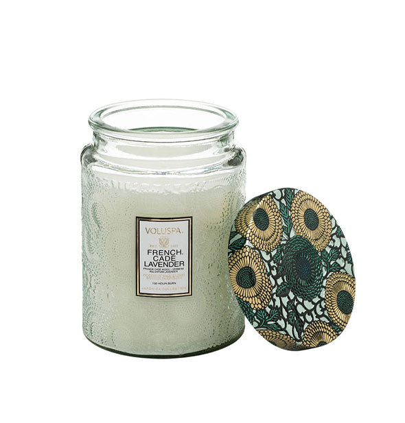 Light green embossed glass Voluspa French Cade Lavender candle jar with gold and green floral patterned tin lid removed and propped up alongside