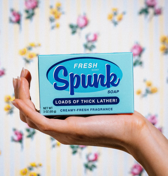 Model's hand holds a blue box of Fresh Scent Spunk Soap up in front of a floral backdrop
