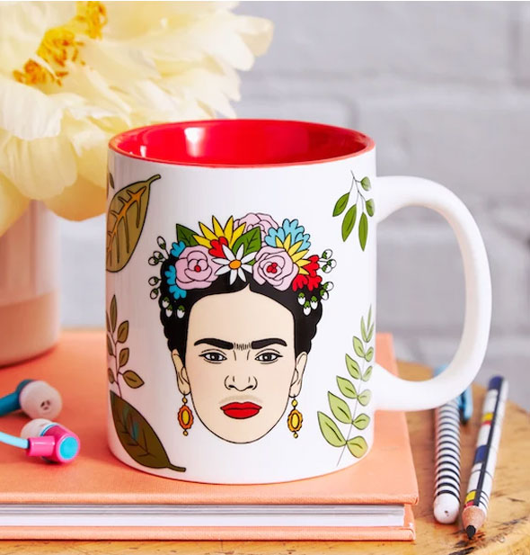 Frida Kahlo mug on top of a book next to pencils, earbuds, and a flower vase