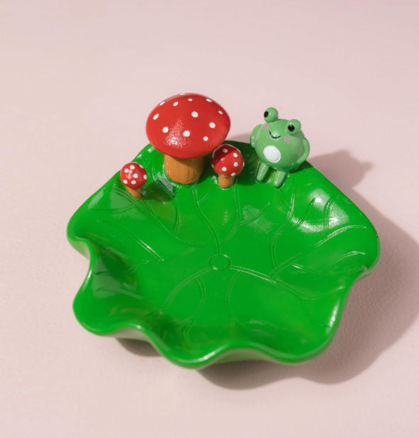 Green lily pad ashtray with curled edges features green frog and red and white spotted toadstools accents