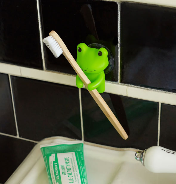 A wooden toothbrush rests between the arms of a little green frog attached to black bathroom tile with a suction cup