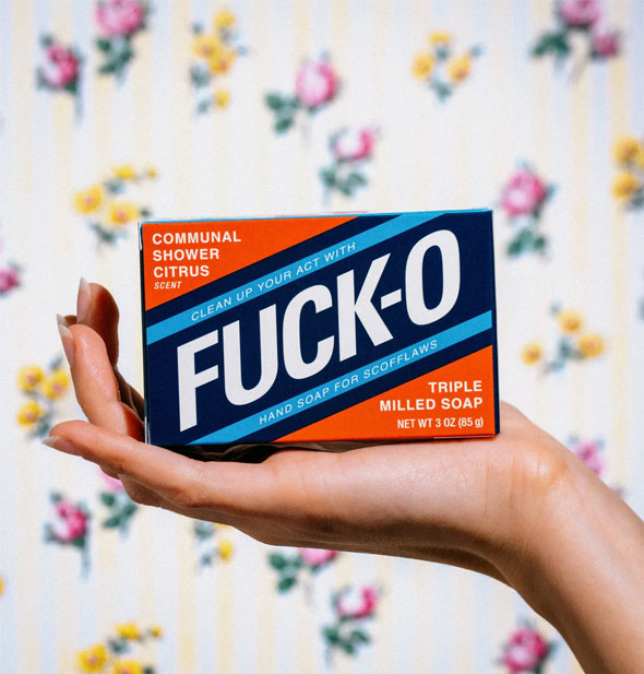 Model's hand holds an orange, blue, and white Fuck-O Triple Milled Soap box up in front of a floral backdrop