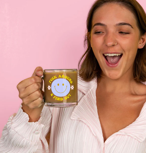 Smiling model holds a Fuck Around & Find Out glass mug filled with a coffee-like beverage by its handle in front of a pink backdrop