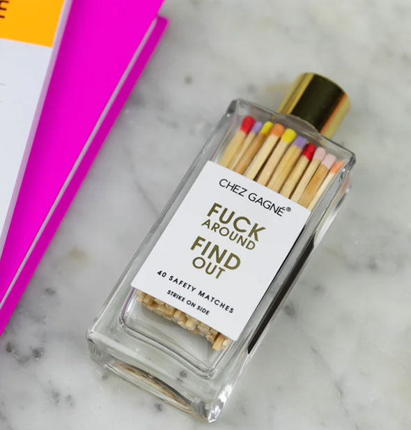 Fuck Around Find Out safety match bottle rests on its side on a marble surface next to bright pink, yellow, and white notebooks