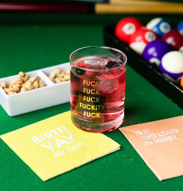 Fuck Fuck Fuck Fuckity Fuck rocks glass on billiard table with printed napkins, pool cues and balls, and dish of nuts is filled with an iced pink-red beverage