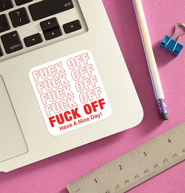 Fuck Off Have a Nice Day! sticker is applied to a laptop staged with pencil, ruler, and blue binder clip on a pink surface