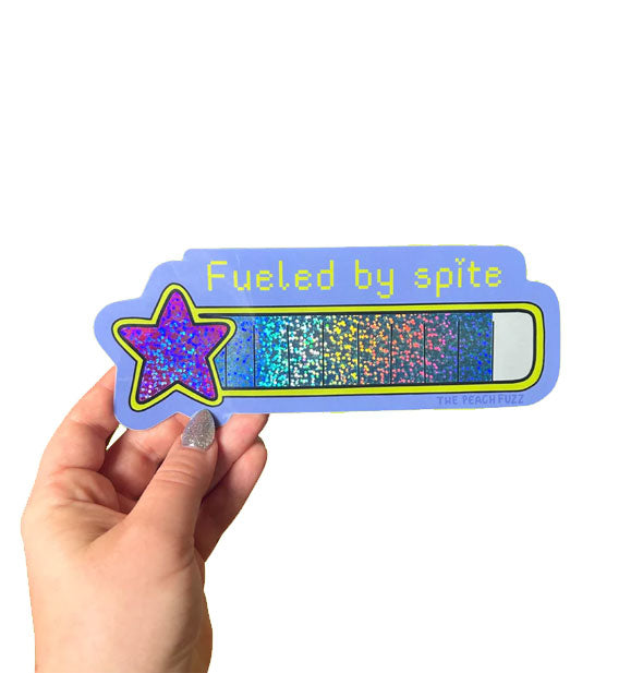 Model's hand holds a sticker with glittery battery level gauge with a star and the end and says, "Fueled by spite" in yellow pixelated lettering